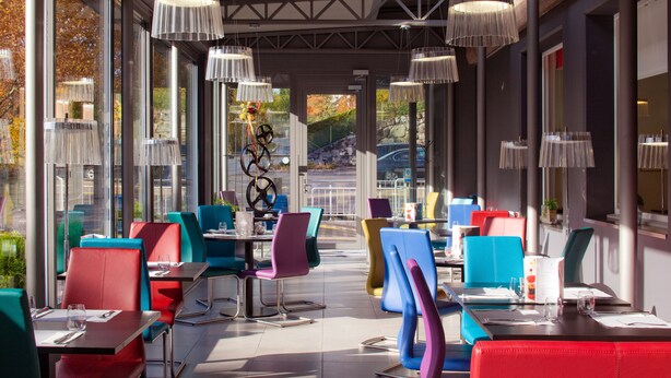 Un área de comedor al aire libre con sillas y mesas de colores, ventanas amplias y lámparas colgantes.