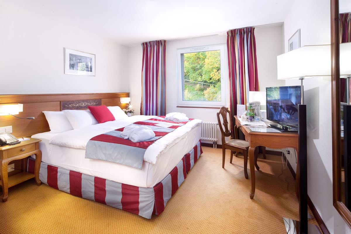 Une chambre d’hôtel avec un grand lit, un bureau avec un ordinateur, une chaise, une télévision et une fenêtre avec des rideaux.