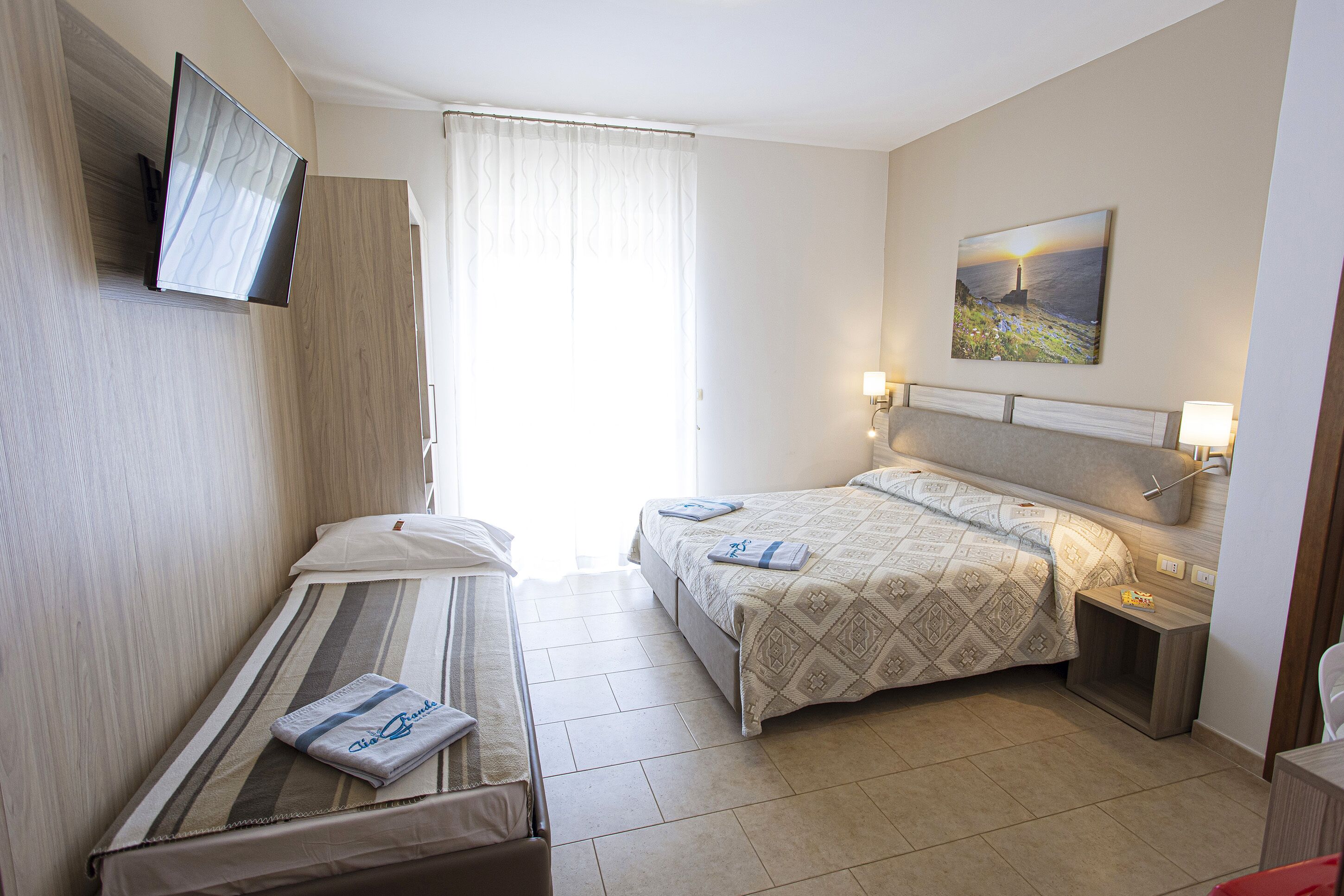A hotel room with two beds, a TV mounted on the wall, a painting above the headboard, and a window with curtains.