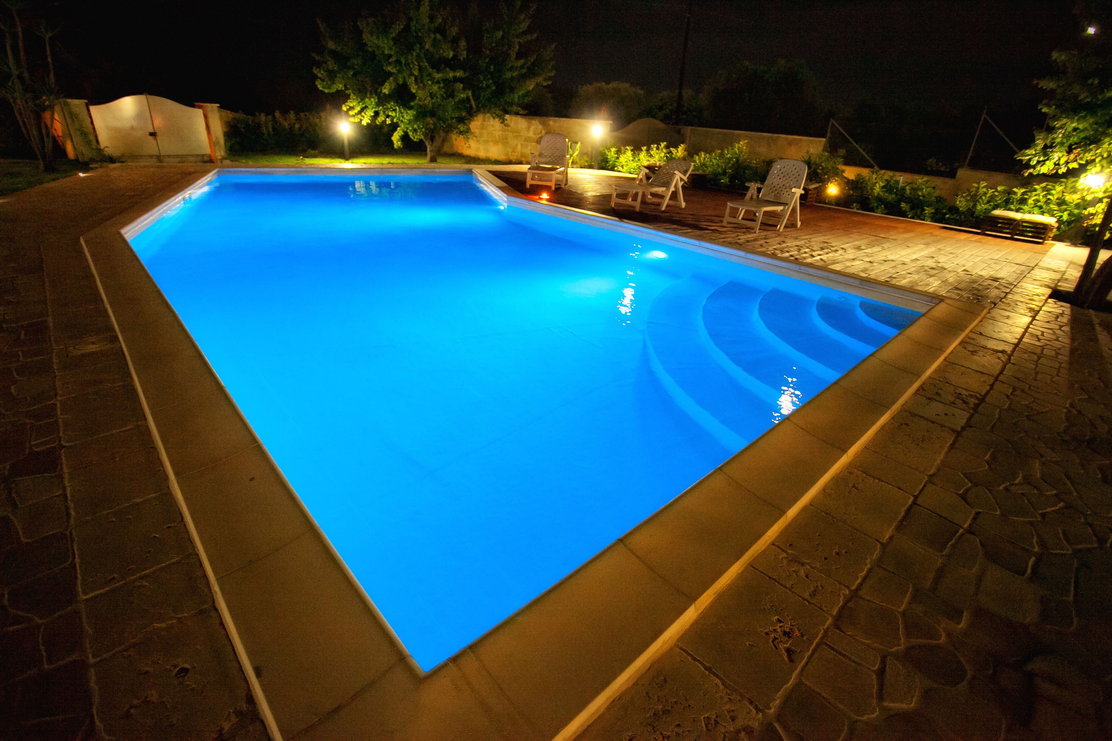 Una piscina rectangular con superficie de agua azul cristalina, rodeada por un patio de piedra con algunas sillas de descanso y una mesita.