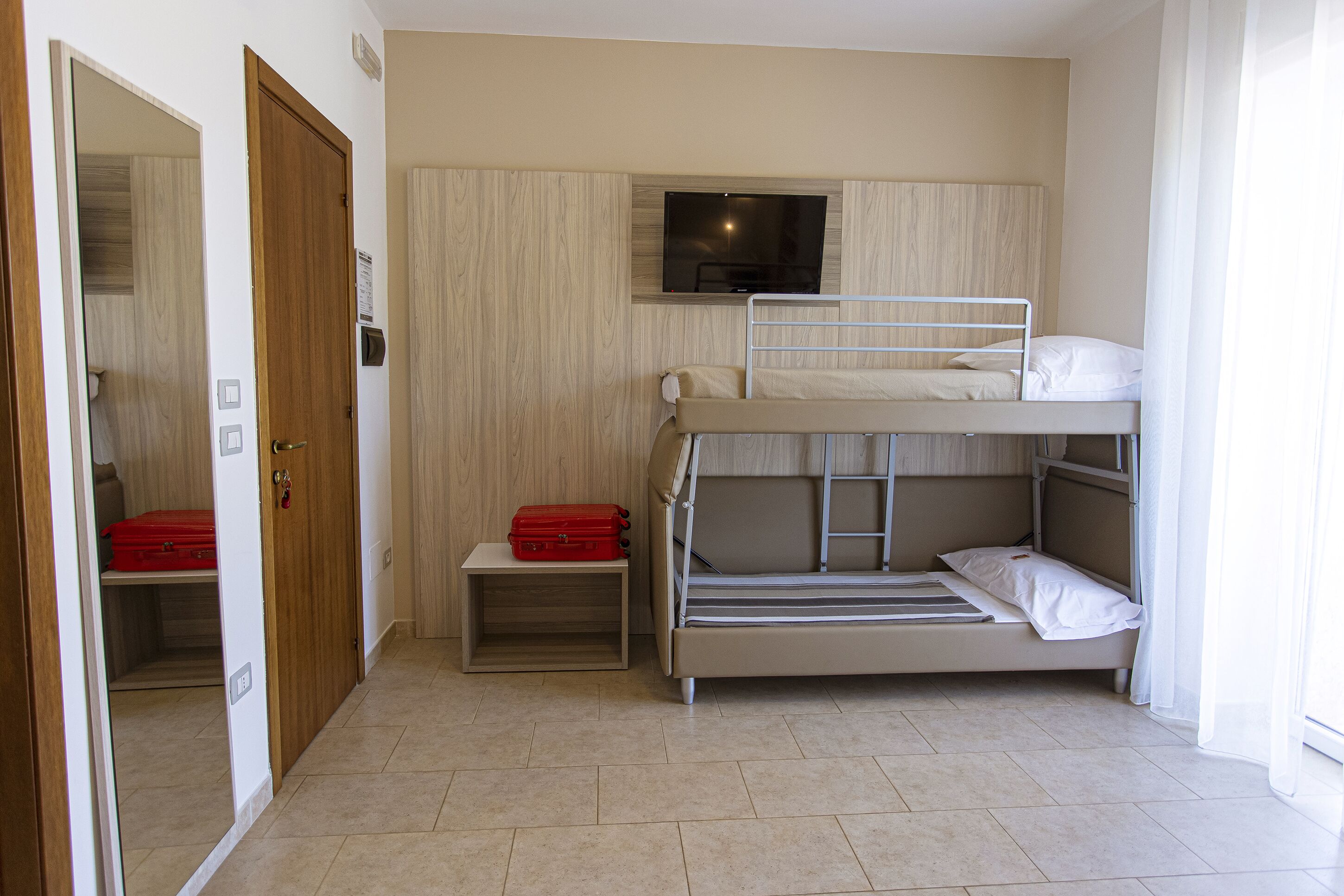 Een slaapkamer met een stapelbed en een televisie erboven, een rode koffer op een klein tafeltje, en een deur die leidt naar een inloopkast.