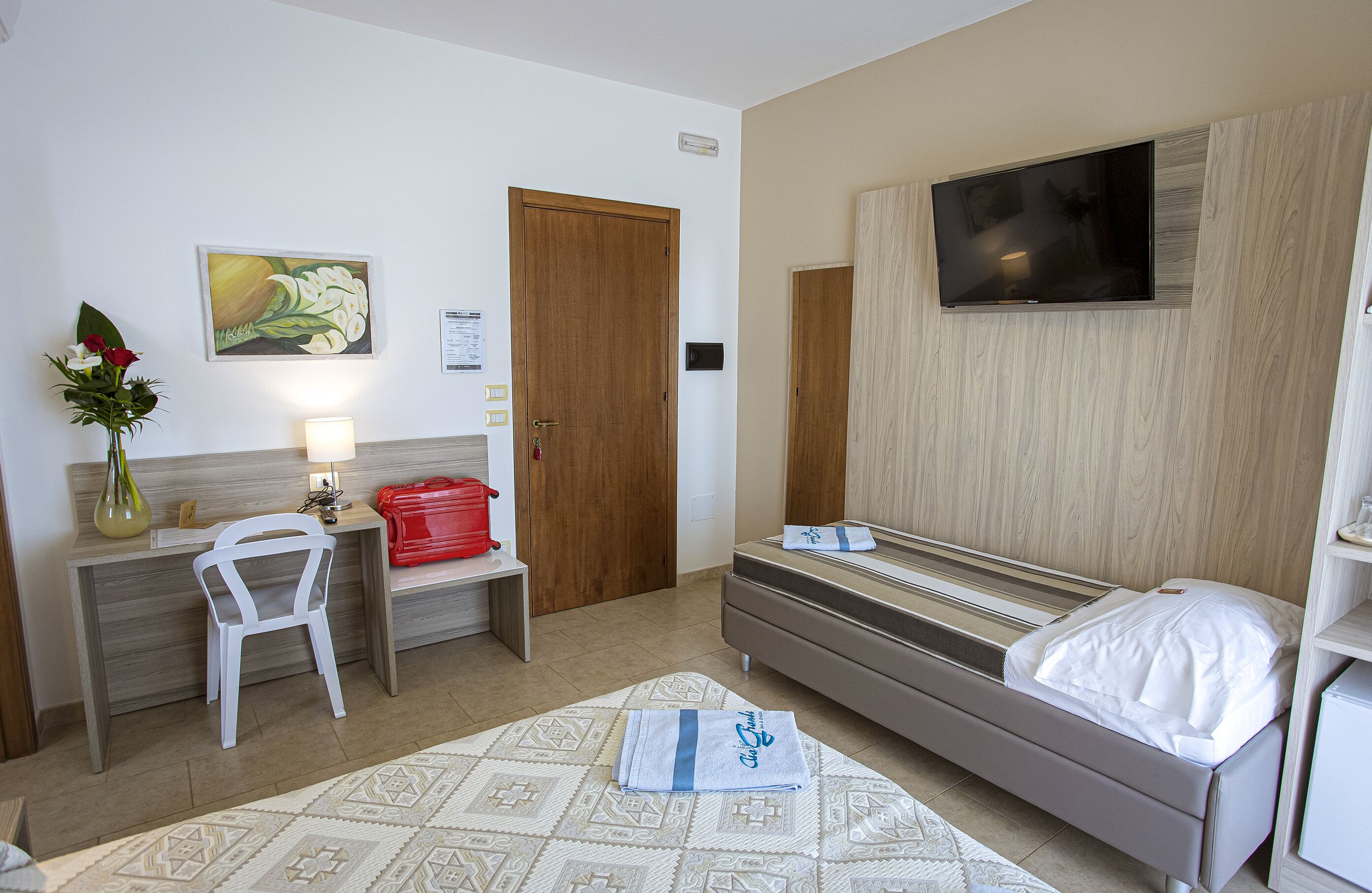 A hotel room with a bed, a desk with a chair, a TV mounted on the wall, and a painting on the wall.