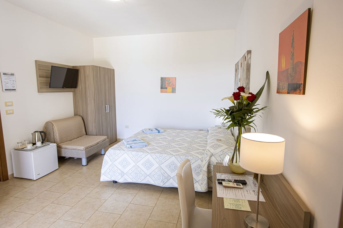 A hotel room with a bed, a desk with a lamp, a chair, a small sofa, a TV mounted on the wall, and a potted plant.