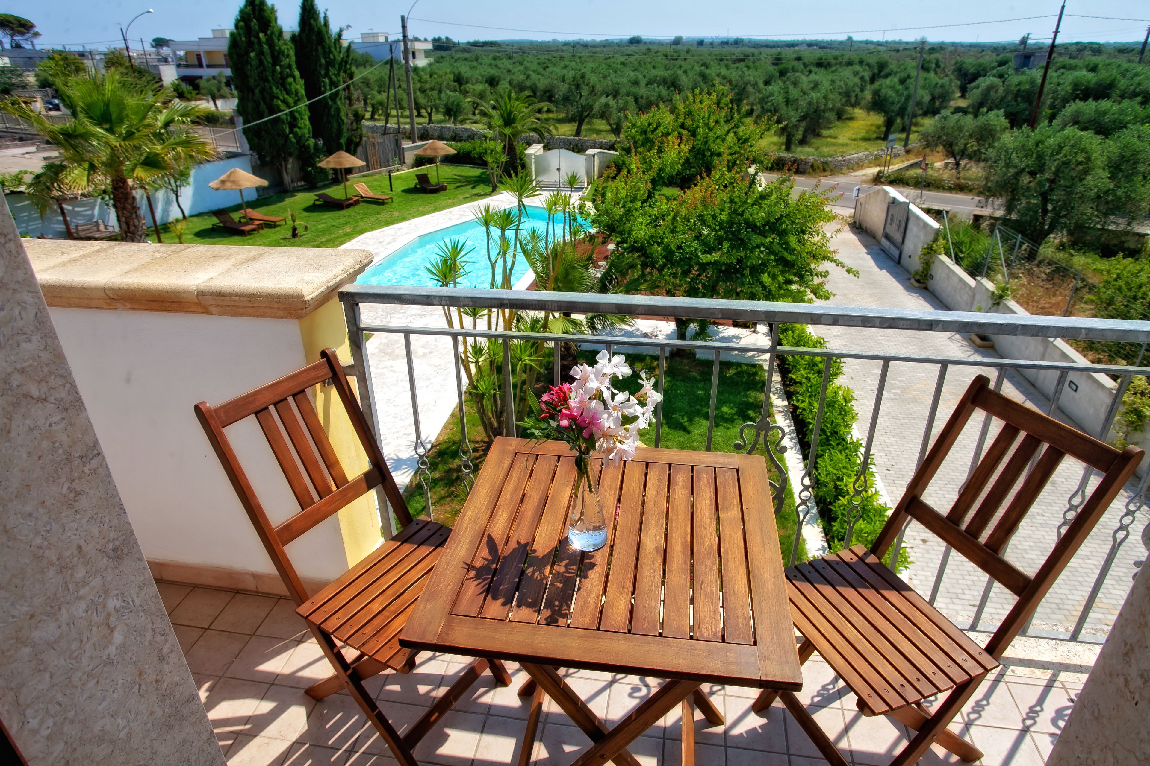 Un balcón con mesa y sillas de madera, un florero con flores y vistas a una piscina y vegetación.