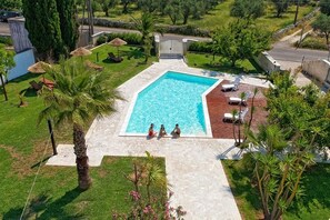 Una piscina con tres personas sentadas cerca, rodeada de palmeras y un área de patio de ladrillo.