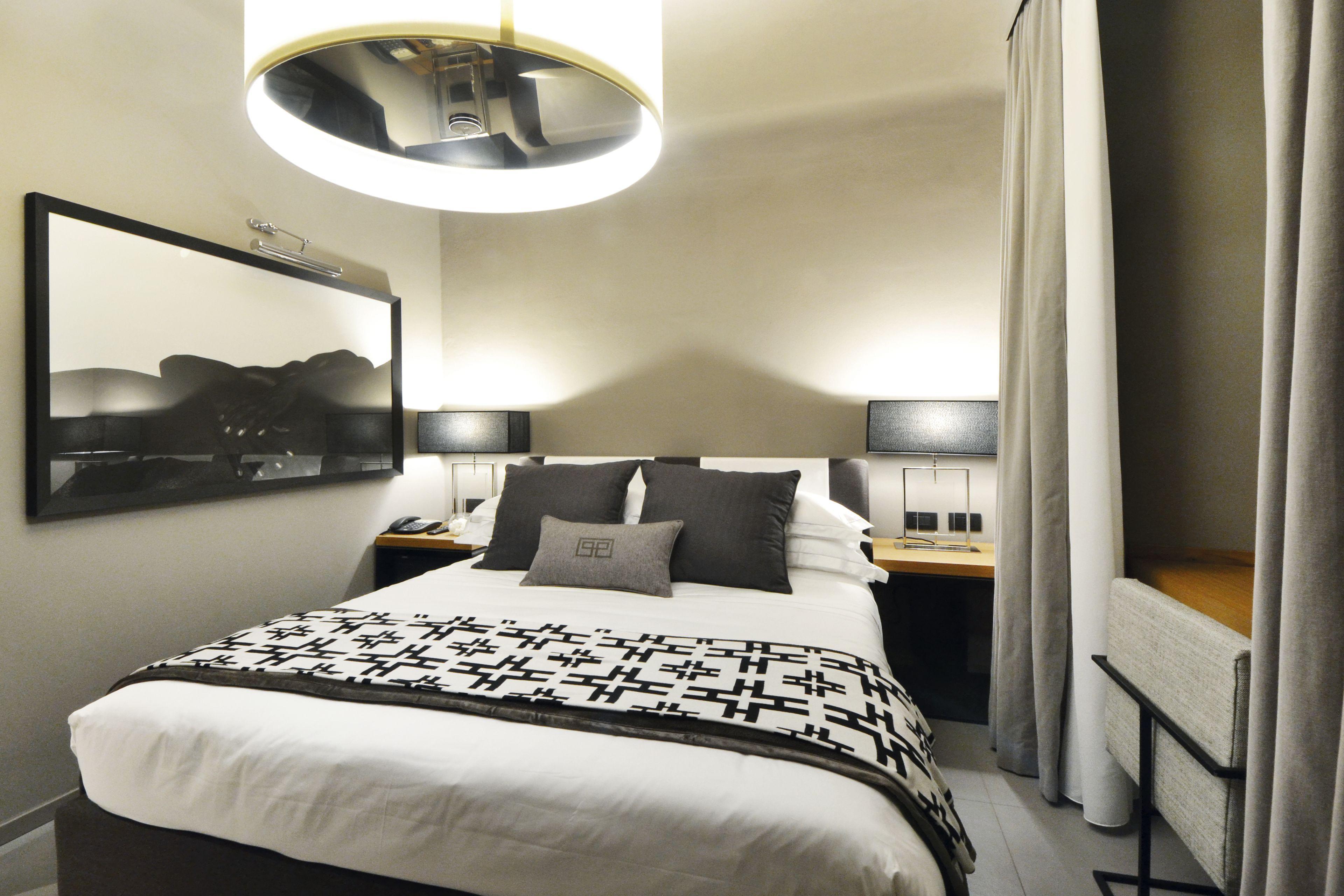 A hotel room with a bed, bedside tables, a desk, a lamp, a mirror, and curtains.