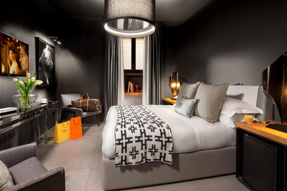 Una habitación de hotel moderna con una cama grande, mesitas de noche, un escritorio con silla, un ventanal con cortinas y un televisor de pared.