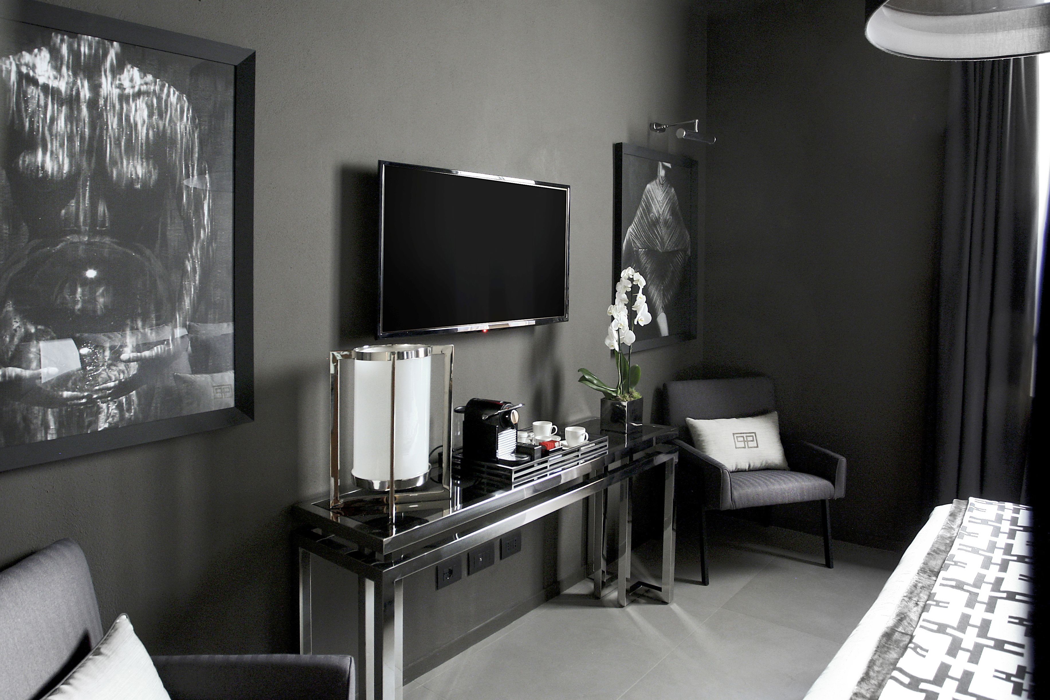 Una habitación de hotel moderna con un televisor de pantalla plana grande montado en la pared, una cafetera sobre una mesita consola, un sillón cómodo y una cama con colcha estampada.