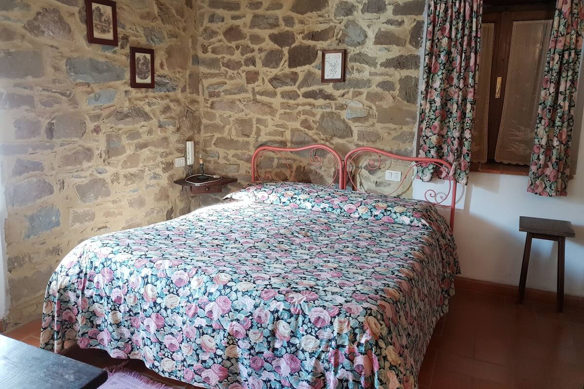 A bedroom with a stone wall, a floral bedspread, a metal bed frame, a bedside table with a lamp, and a window with floral curtains.