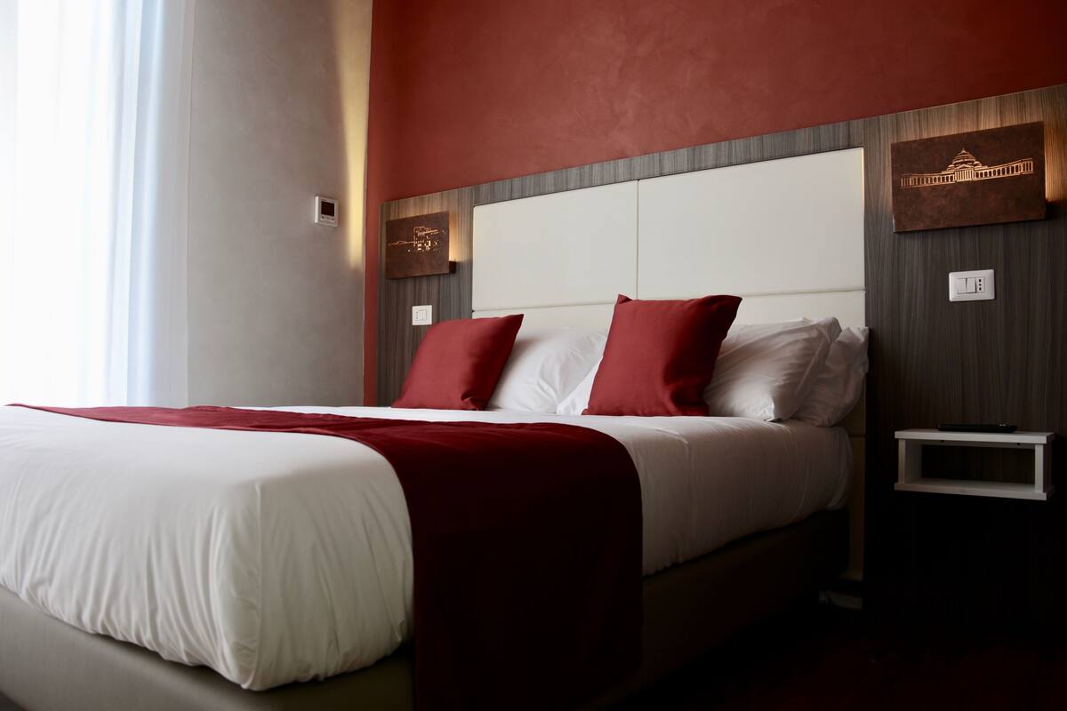 Ein Hotelzimmer mit einem großen Bett, rot-weißer Bettwäsche, einem Kopfteil mit integrierter Beleuchtung und einem kleinen Nachttisch.