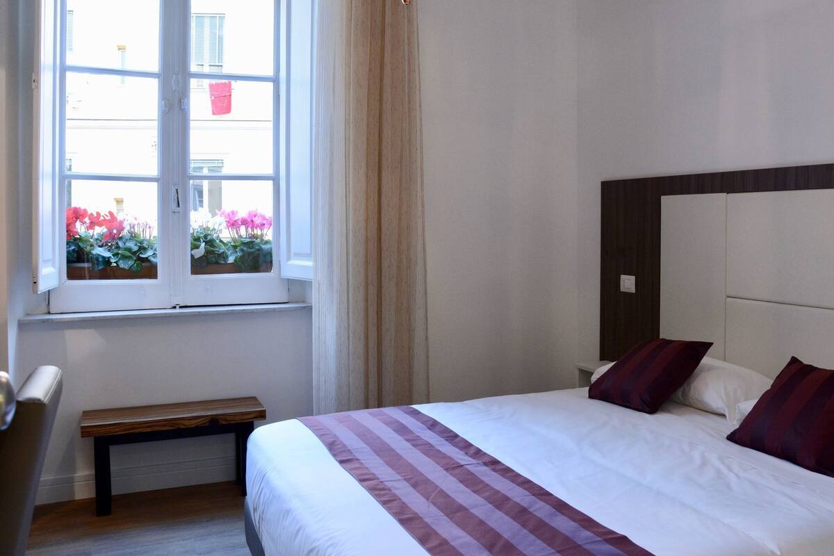 Ein Hotelzimmer mit einem großen Bett, einer hölzernen Bank, einem Fenster mit Blick auf Blumen und einer schlichten Wand.