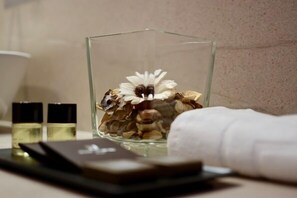 Un recipiente de vidrio con flores secas y frutos secos, una toalla blanca y dos botellas pequeñas sobre una superficie reflectante.