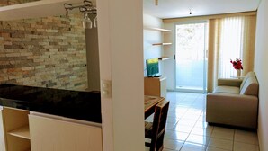 Eine moderne Wohnung mit Küche, die einen schwarzen Arbeitsbereich, eine Steinwand und einen Essbereich mit Tisch und Stühlen bietet. Es gibt ein Wohnzimmer mit einer Couch und einem Fernseher. Die Wohnung verfügt über einen Balkon mit einer Schiebetür und Blick ins Freie.