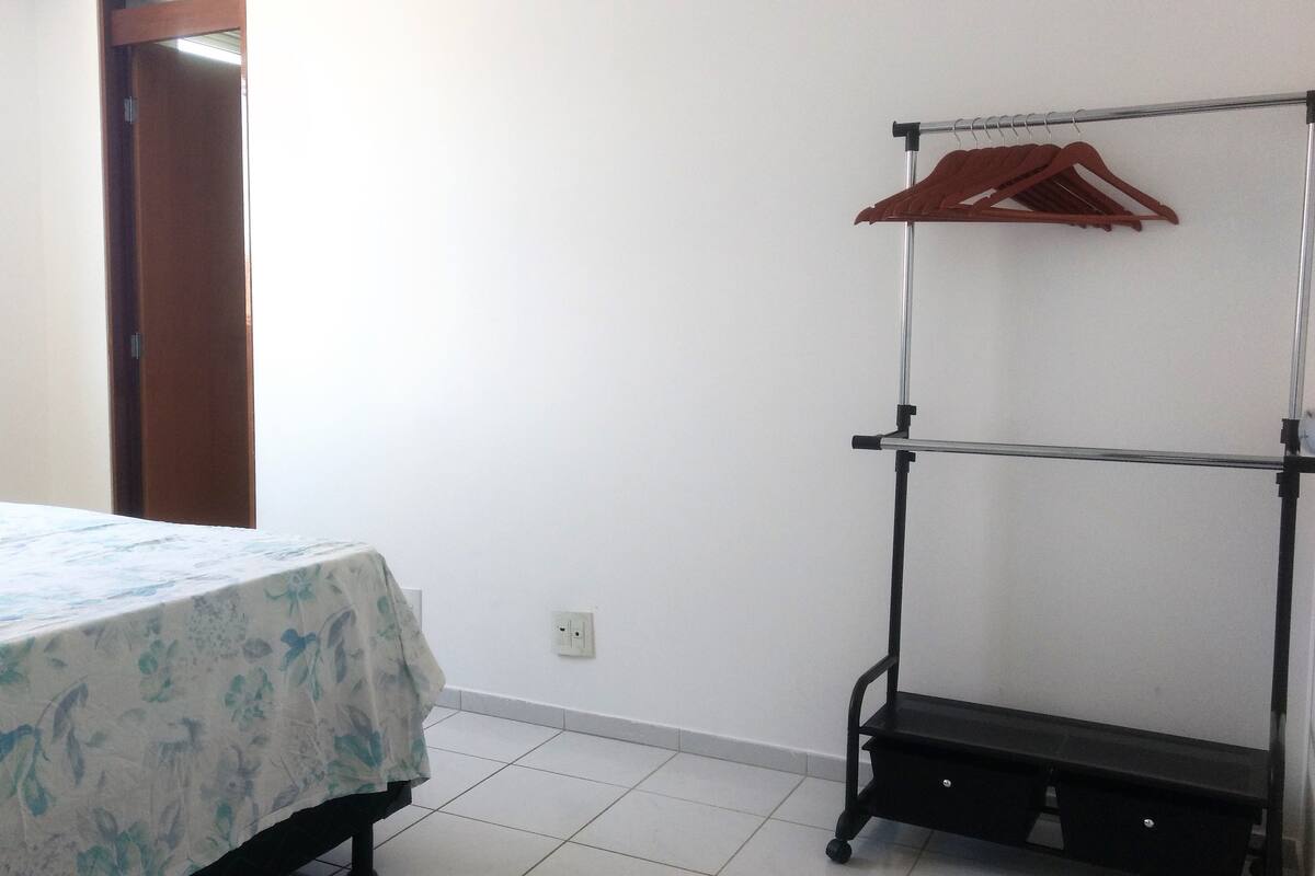 Una habitación con una cama cubierta con una colcha de estampado floral, un perchero metálico con un perchero marrón, y una pared blanca con un enchufe.