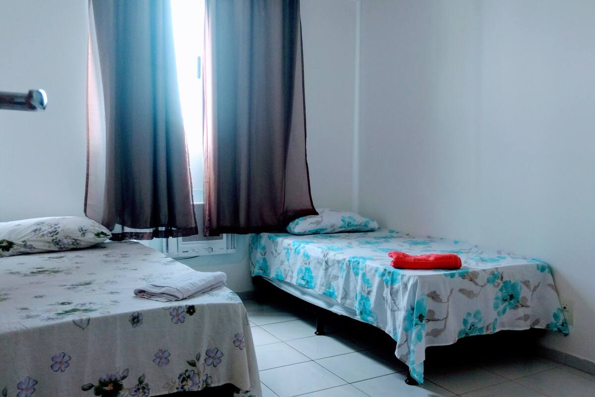Una habitación con dos camas, una con colcha estampada de flores y la otra con colcha azul estampada de flores. Hay un calentador en la pared entre las camas, y un objeto rojo sobre la cama con la colcha azul estampada de flores.