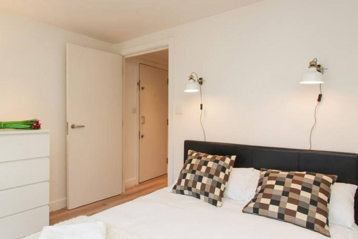 A bedroom with a bed, two bedside lamps, a dresser, and a door leading to another room.