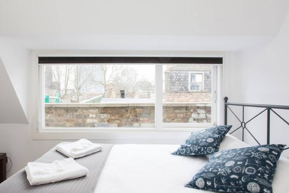 A bedroom with a large window, a bed with white and patterned pillows, a bedside table with a TV and books, and a view of a stone wall and trees outside.