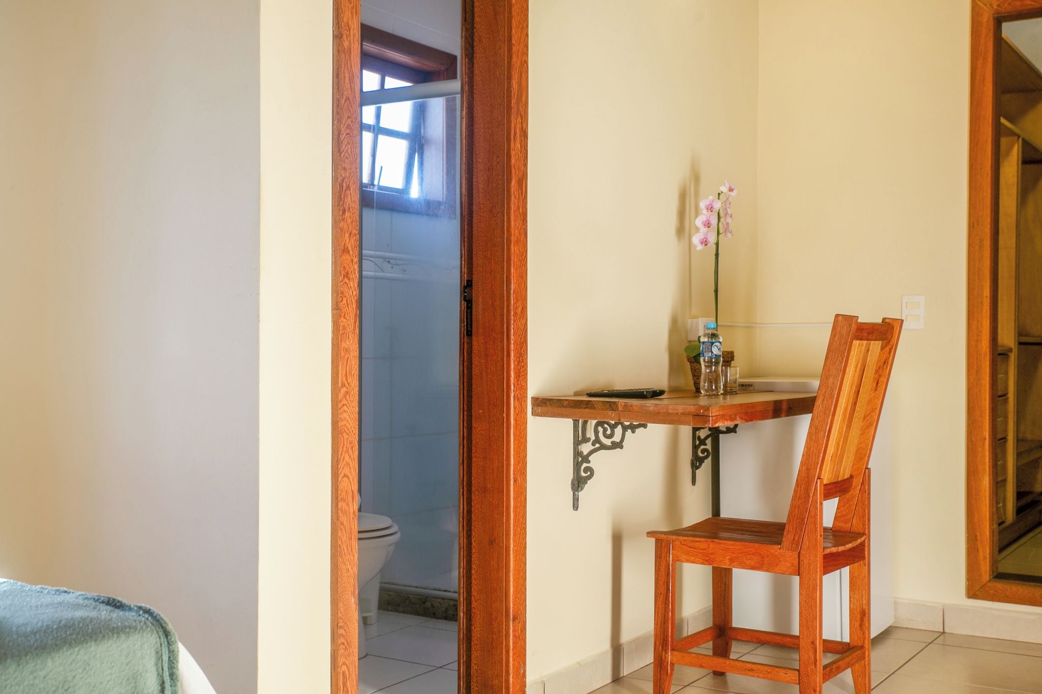 A bathroom with a toilet, a shower, and a small table with a chair. There is a window with natural light and a door leading to another room.