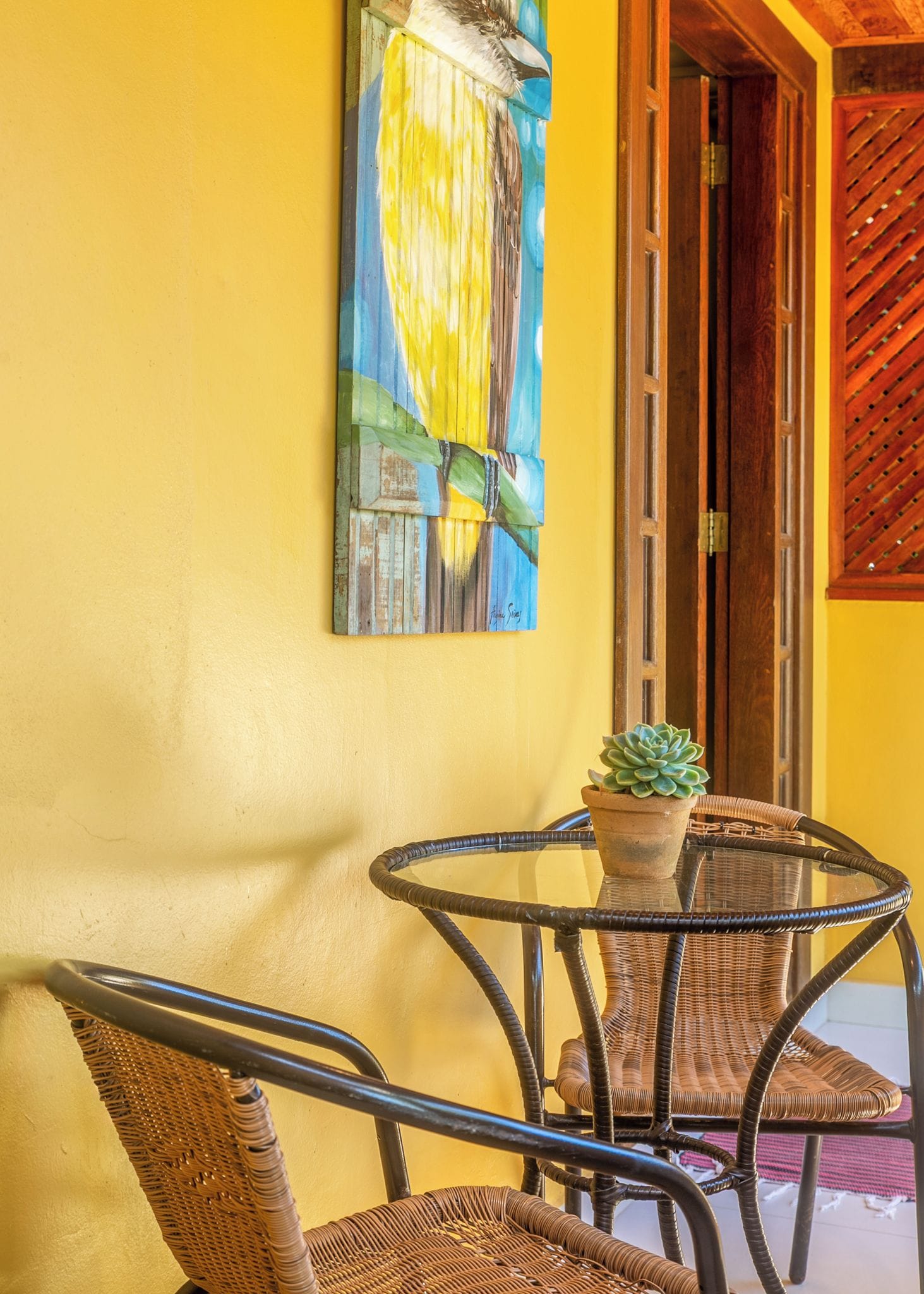 A yellow wall with a painting, a wicker chair, a potted plant, and a wooden door.