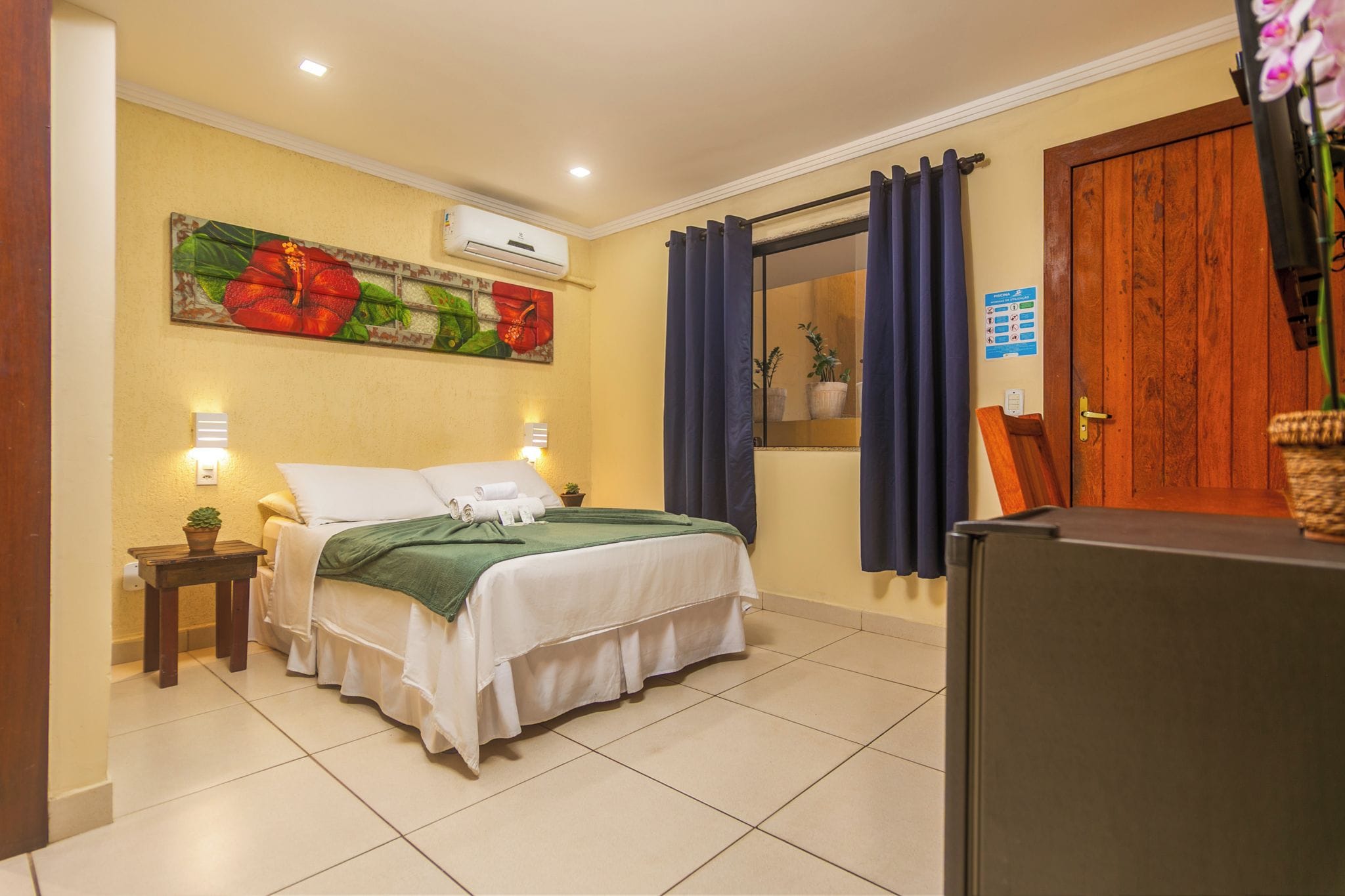 A hotel room with a bed, a small table with a plant, a window with curtains, and a door with a sign.