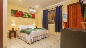 A hotel room with a bed, a small table with a plant, a window with curtains, and a door with a sign.