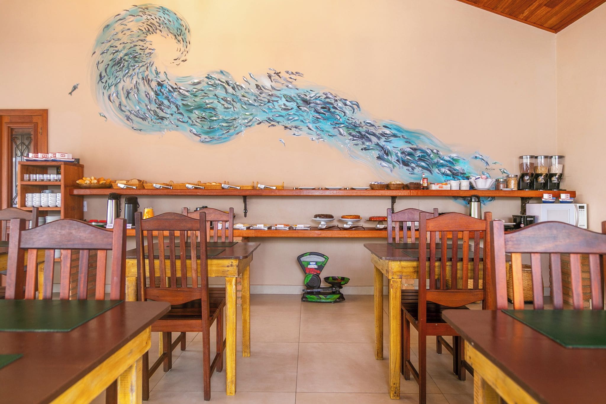 A dining area with wooden chairs and tables, a kitchen counter with various items, and a wall mural of fish.