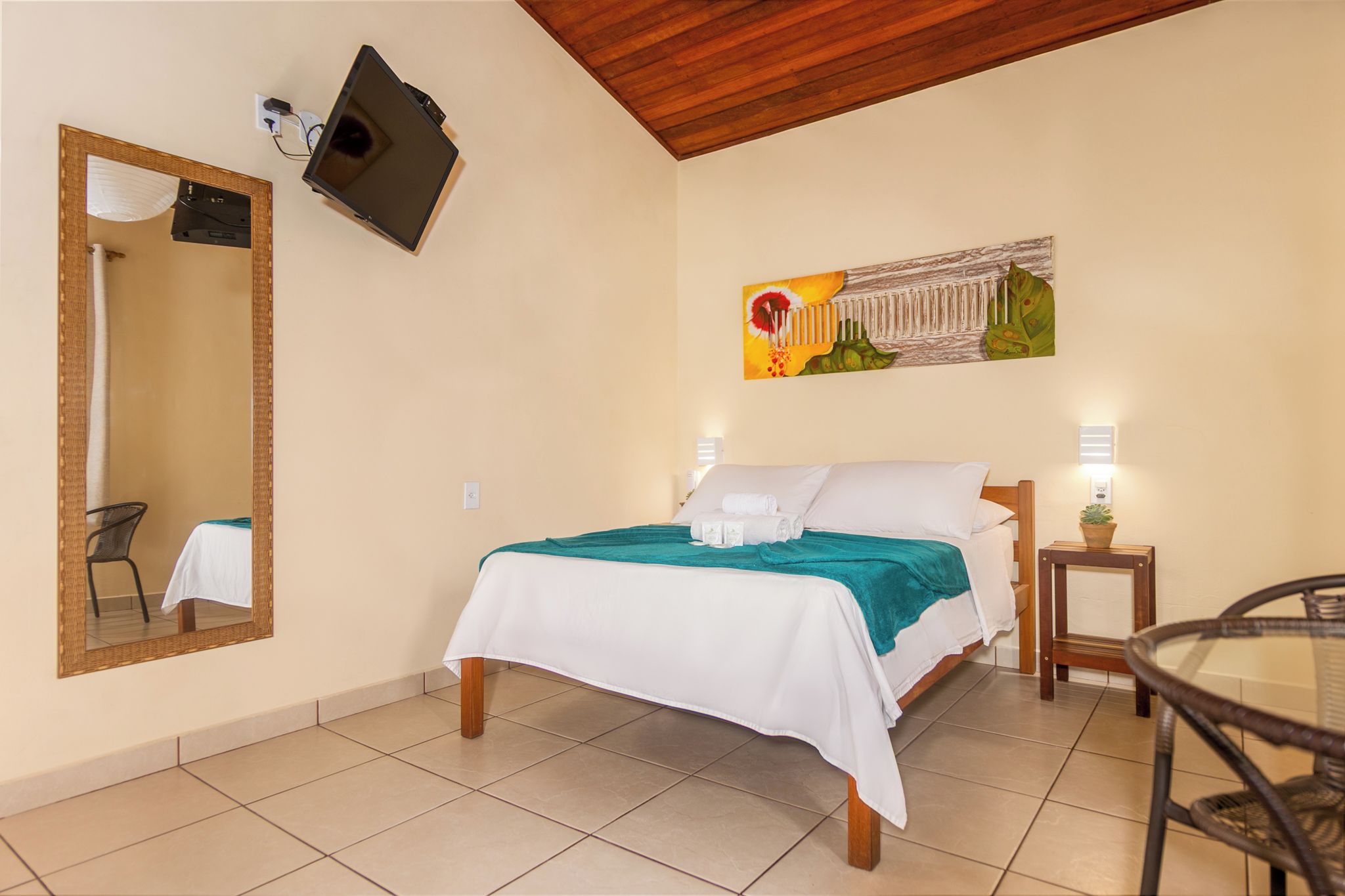 A hotel room with a bed, a chair, a table, a mirror, and a painting on the wall.