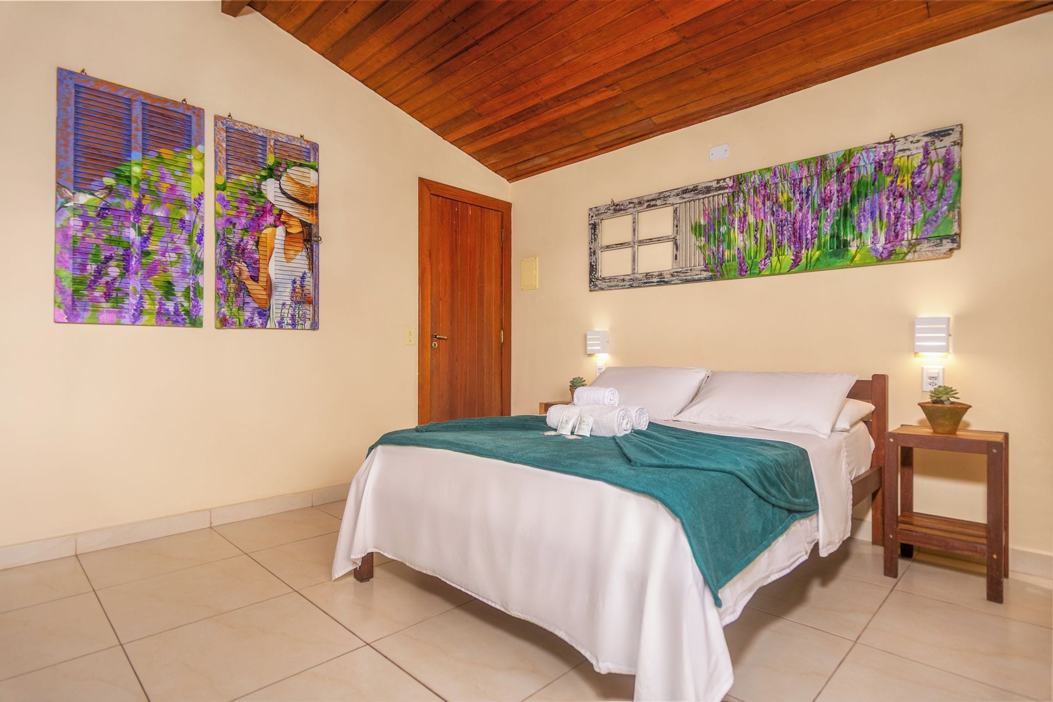 A bedroom with a bed, two wall-mounted paintings, a wooden door, a bedside table with a lamp, and a small potted plant.