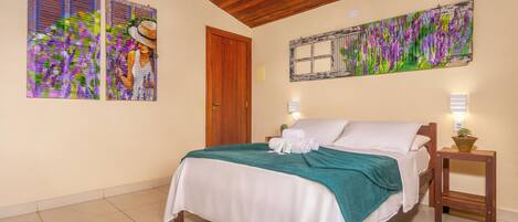 A bedroom with a bed, two wall-mounted paintings, a wooden door, a bedside table with a lamp, and a small potted plant.
