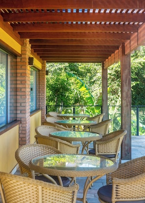 Un espace extérieur aménagé, abrité et doté de mobilier en osier et de tables en verre, offrant une vue sur un jardin luxuriant.