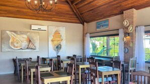 A dining area with wooden tables and chairs, a chandelier, and artwork on the walls. - Pousada Búzios Garden (Búzios)