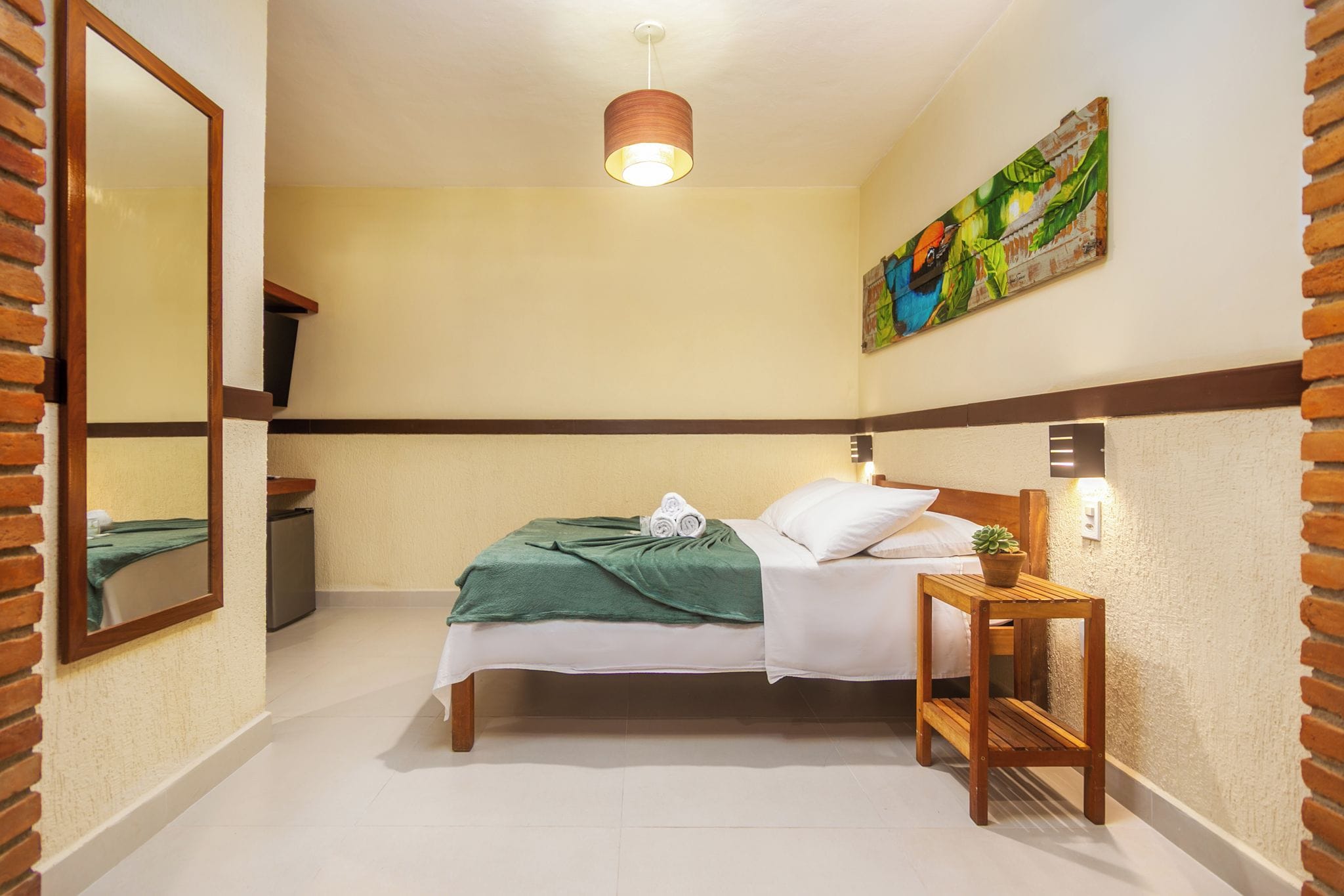 A hotel room with a bed, a bedside table with a plant, a mirror, and a painting on the wall.