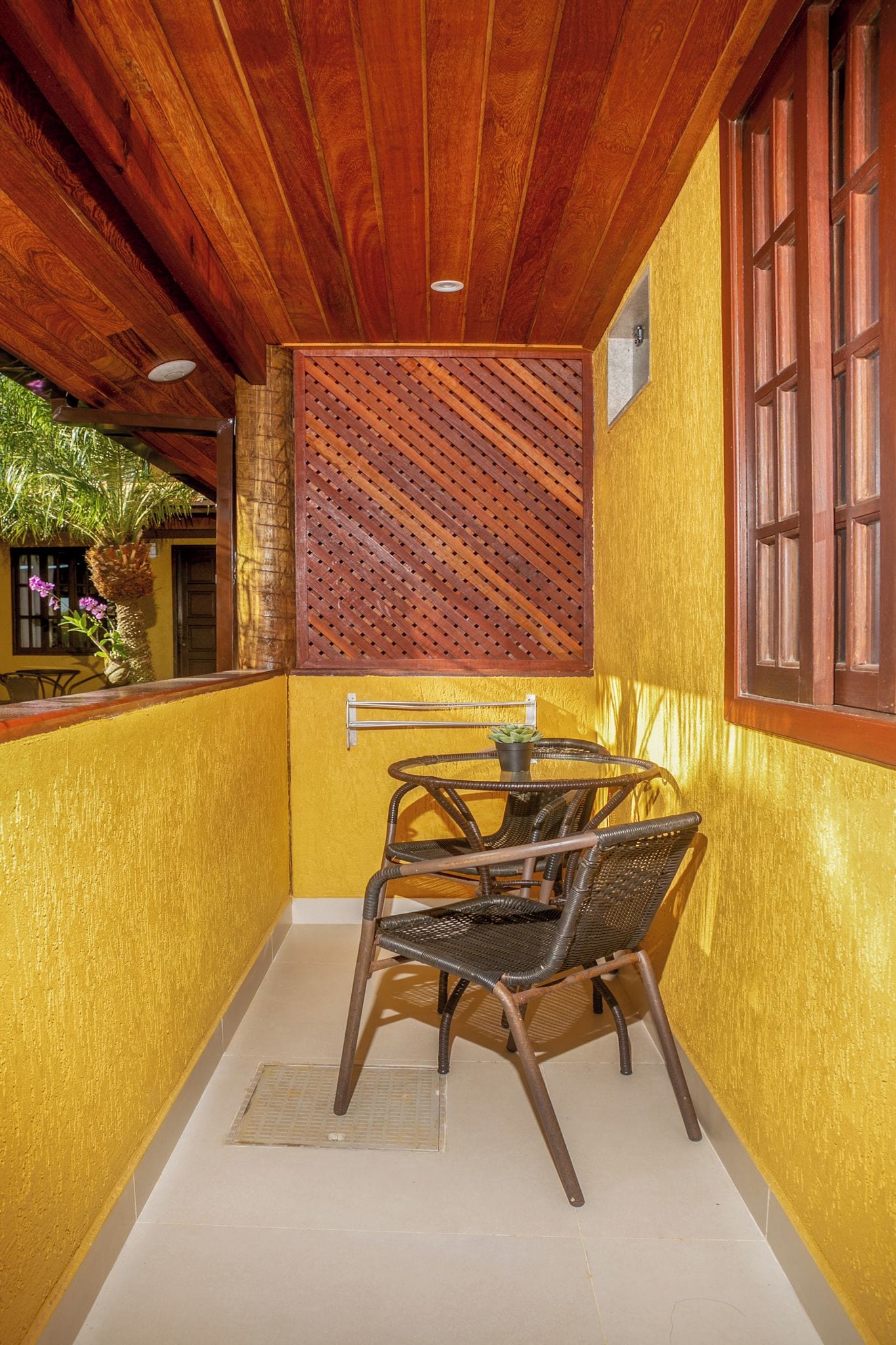 A small, enclosed patio area with a wooden ceiling, a yellow wall, and a window with wooden shutters.