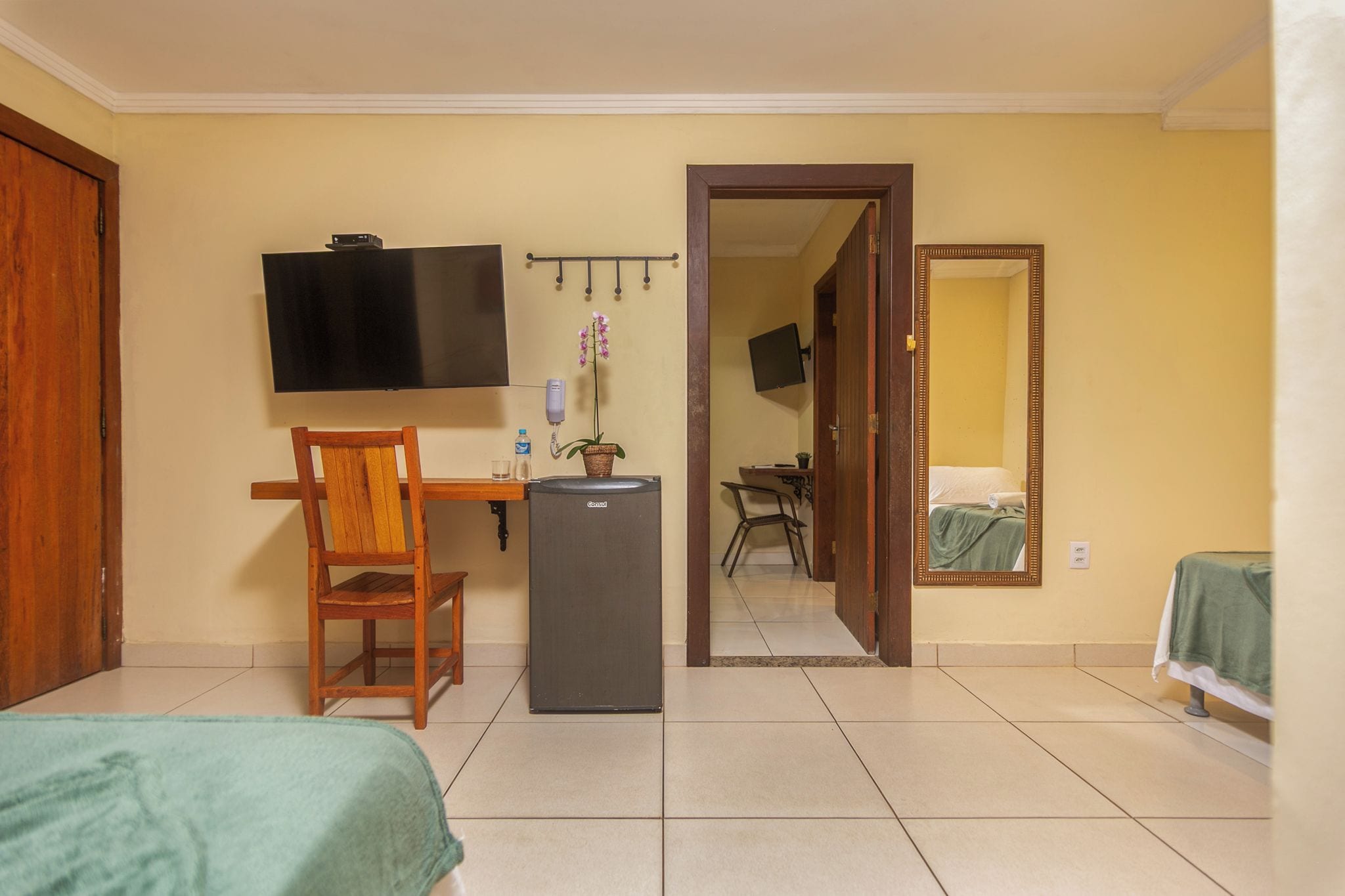 Une chambre d’hôtel comprenant un lit, un bureau avec une chaise, une télévision, un réfrigérateur, un miroir et une porte donnant sur une autre pièce.