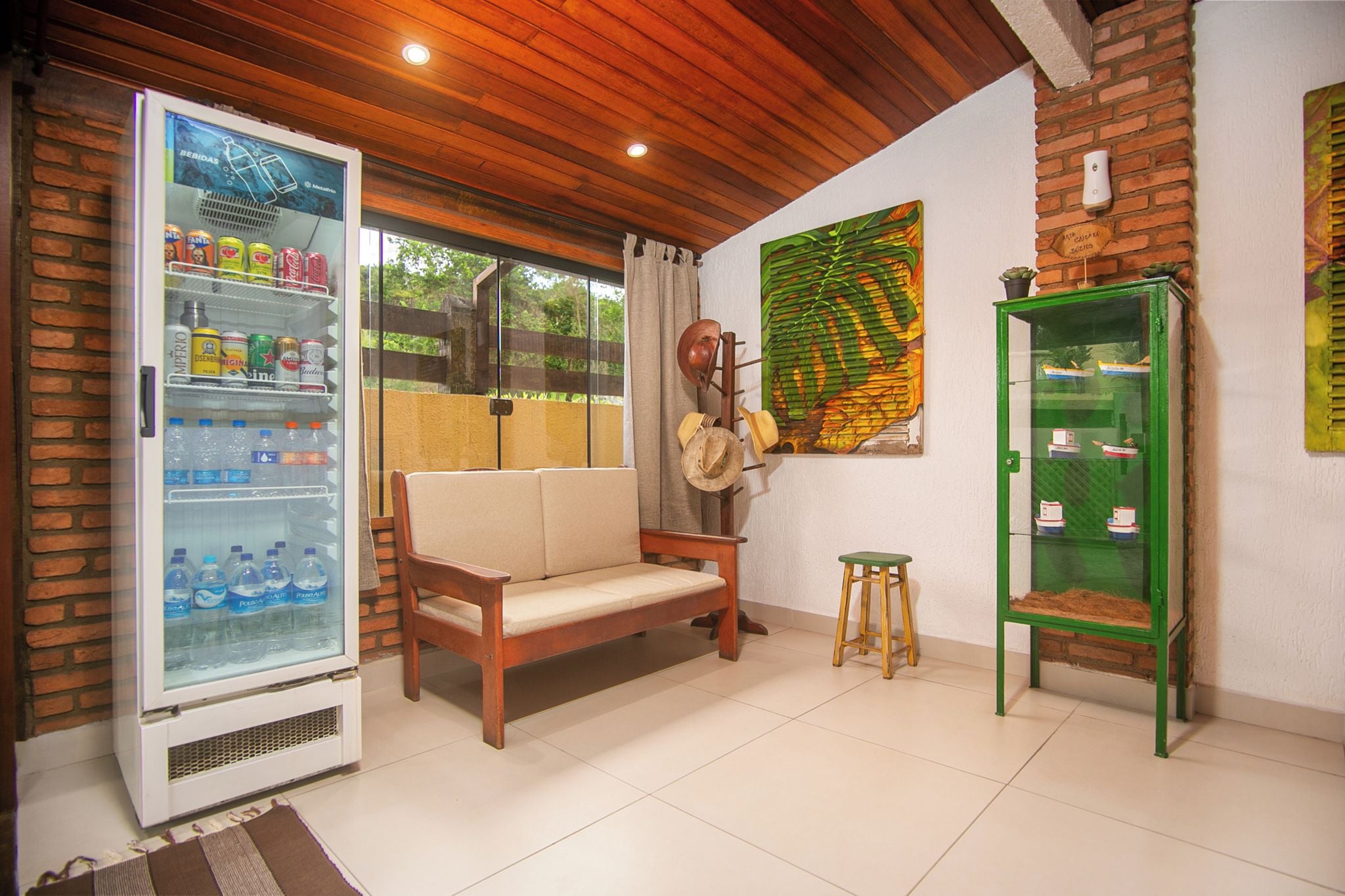 Una habitación con techo de madera, un sofá, un taburete, un refrigerador lleno de bebidas y un armario verde con maquetas de barcos.