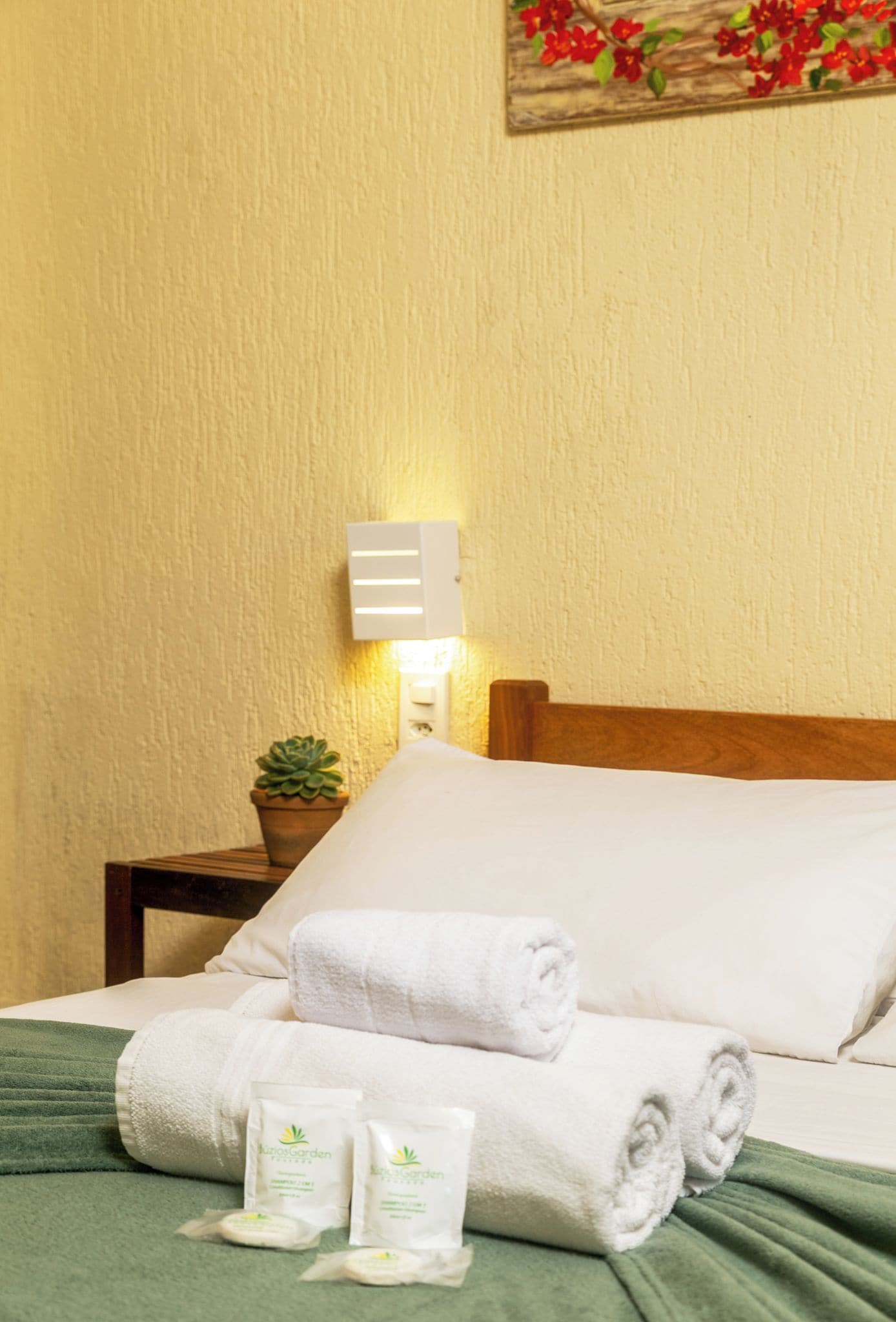 Cama bem arrumada com roupa de cama branca e toalhas enroladas, uma planta na mesinha de cabeceira e luminária fixada na parede.