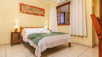 Quarto de hotel com uma cama, mesa de cabeceira com abajur, janela com cortinas e ar condicionado fixado na parede.
