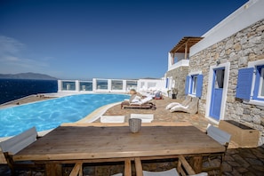 A wooden table with white chairs, a stone wall with blue shutters, and a pool with clear blue water.
