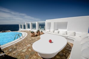 Un espace piscine extérieur moderne, doté d’un sol en pierre, de meubles de salon blancs et offrant une vue sur la mer.
