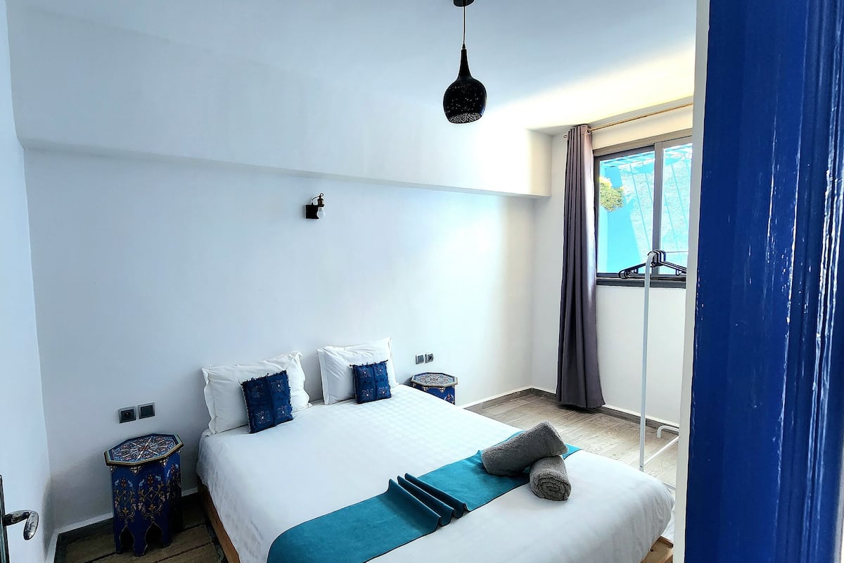 Quarto com cama, mesa de cabeceira, luminária suspensa e janela com cortinas.