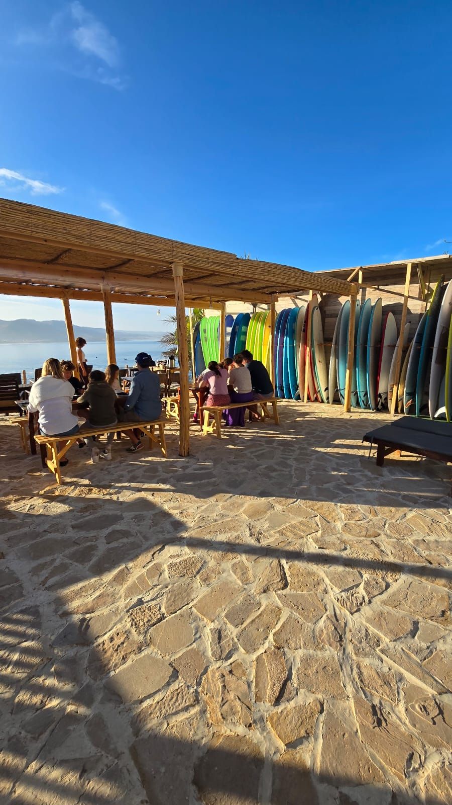 Un café situé en bord de mer, avec des tables et des bancs en bois, entouré de planches de surf aux couleurs vives.