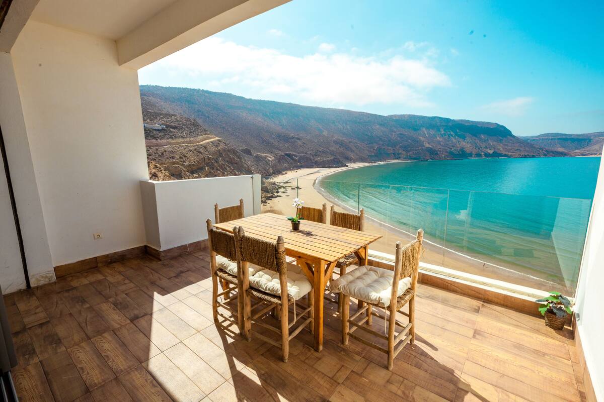 Mesa e cadeiras de madeira numa varanda com vista para uma praia, água turquesa translúcida e colinas ao longe.
