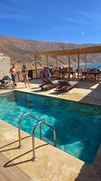 Zona de piscina con agua cristalina, sillones de madera y una pérgola.