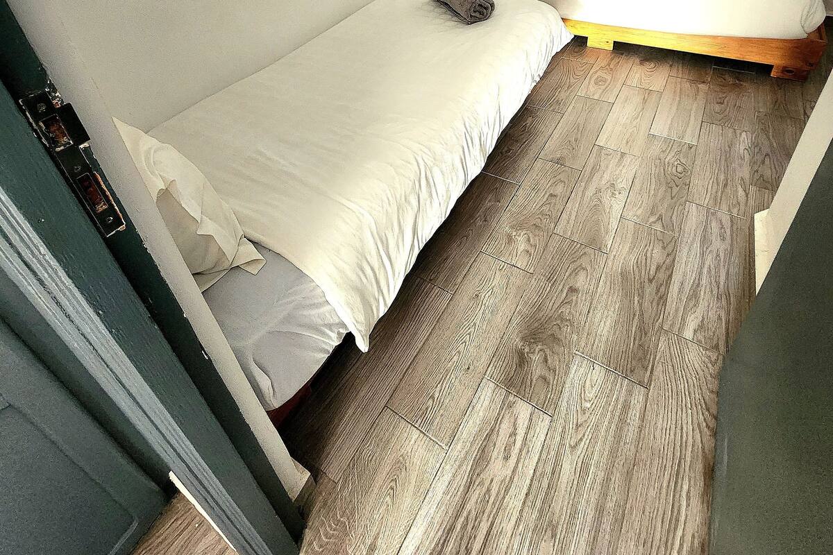 Acomodação pequena, com um quarto e duas camas, piso de madeira e duas luminárias fixadas na parede.