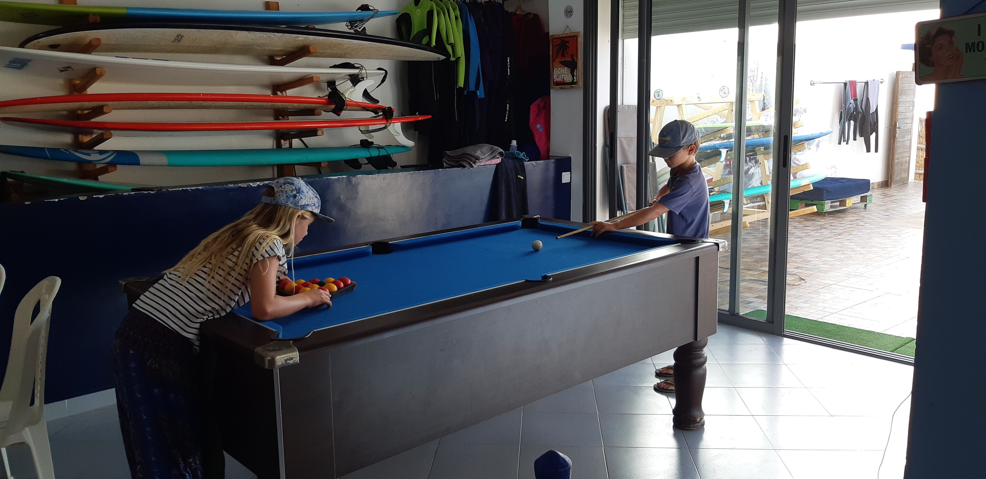 A pool table in a room with surfboards on the wall and a view of the outside.