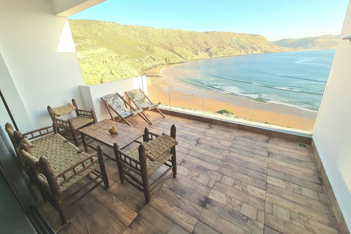 Uma varanda com piso de madeira, uma mesa e duas cadeiras com assentos de vime.