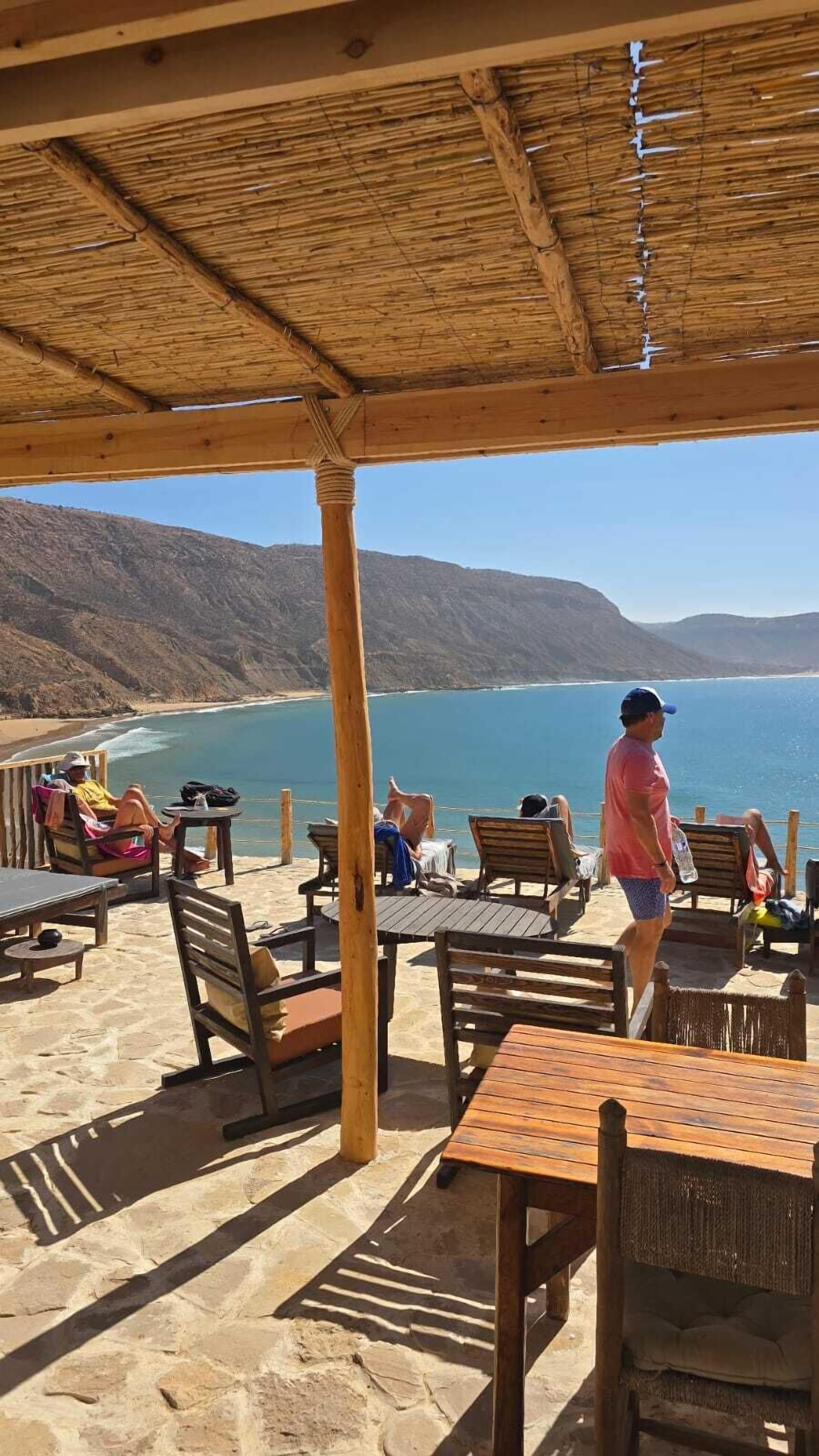 Ein Sitzbereich im Freien mit Holztischen und -stühlen mit Blick auf Strand und Berge.