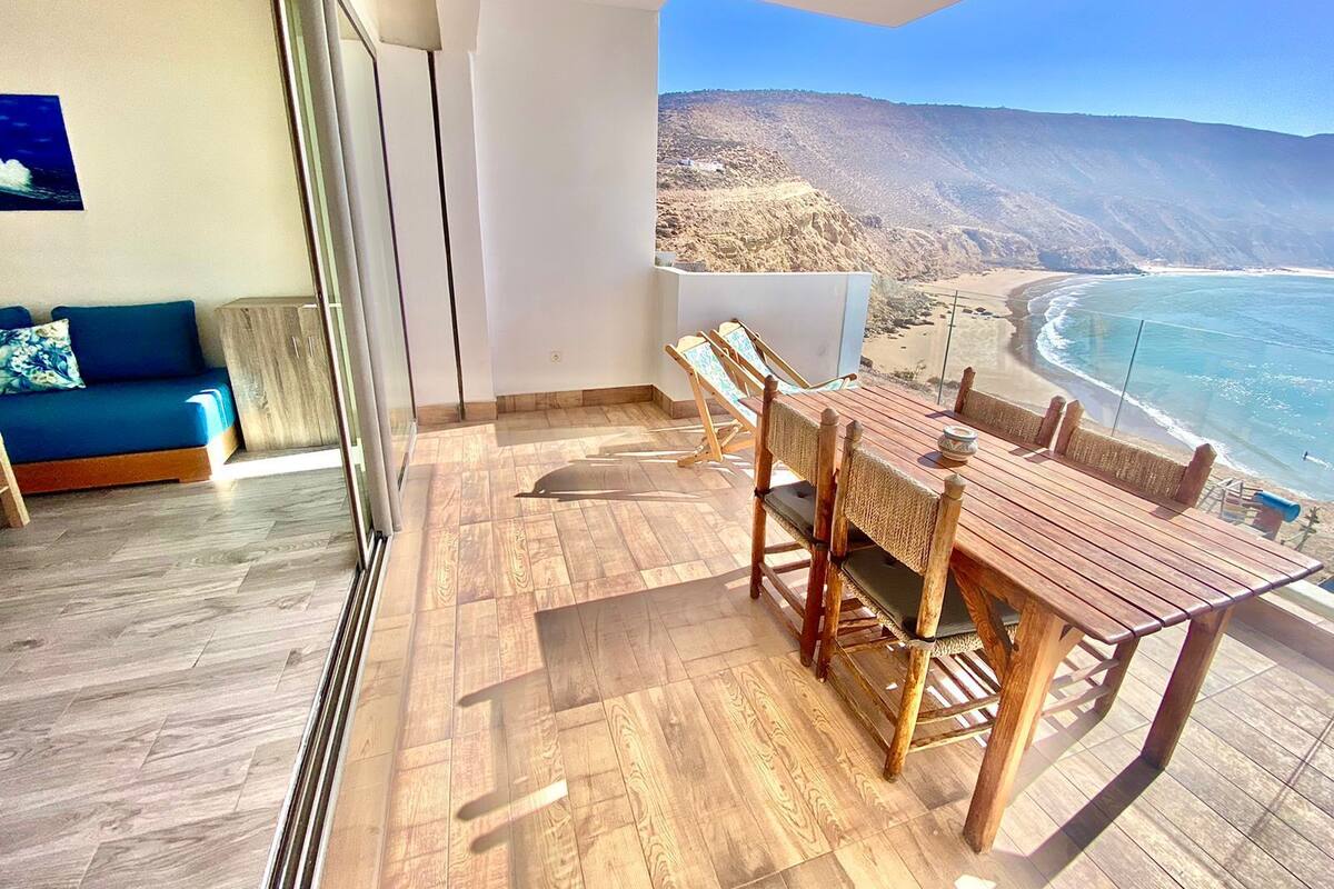 Uma varanda com mesa e cadeiras de madeira, um sofá e vista para a praia e montanhas.