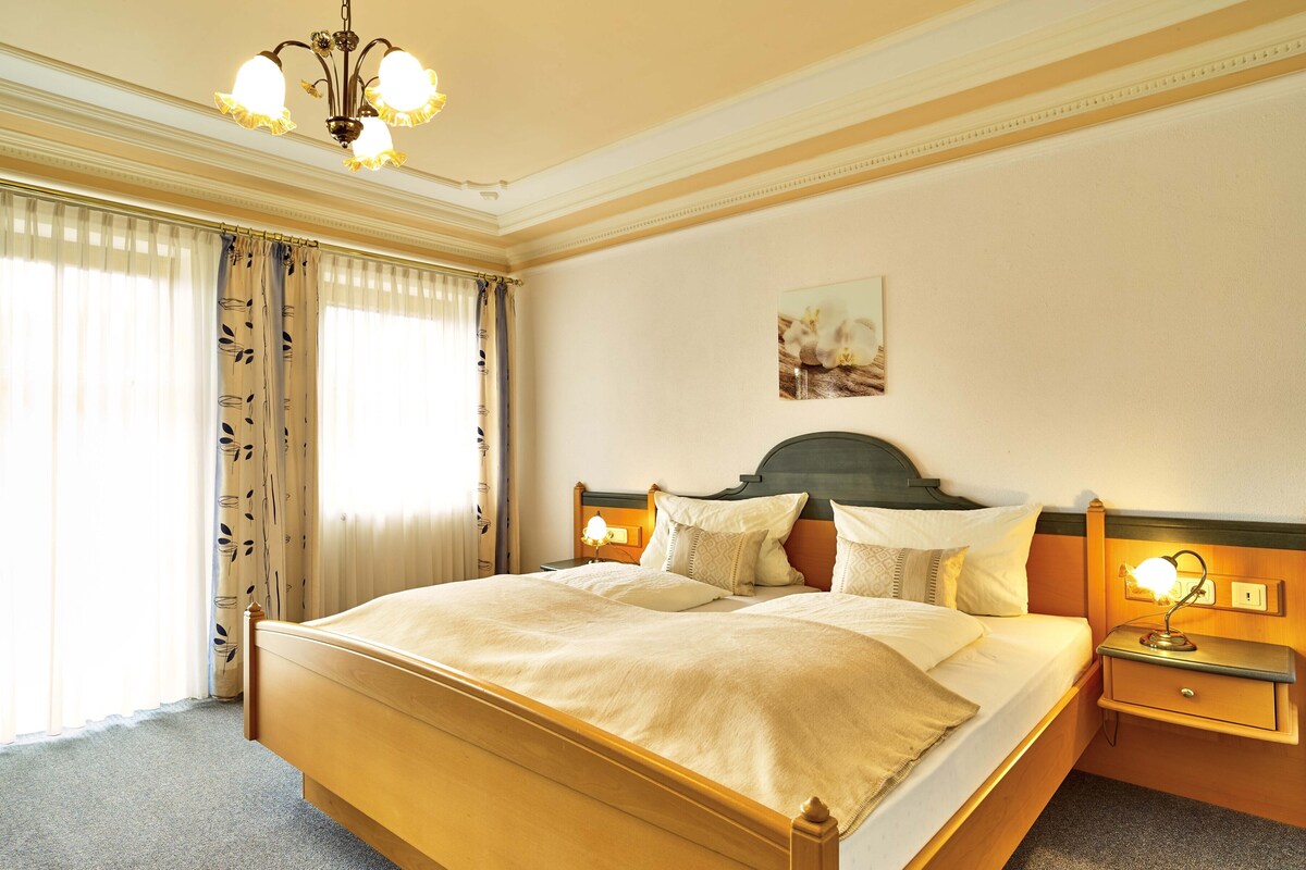 Una camera d'albergo con un letto grande, un lampadario, un comodino con una lampada e un quadro appeso al muro.