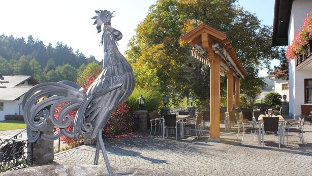 Un patio con una escultura de gallo de metal, mobiliario exterior y una pérgola de madera.